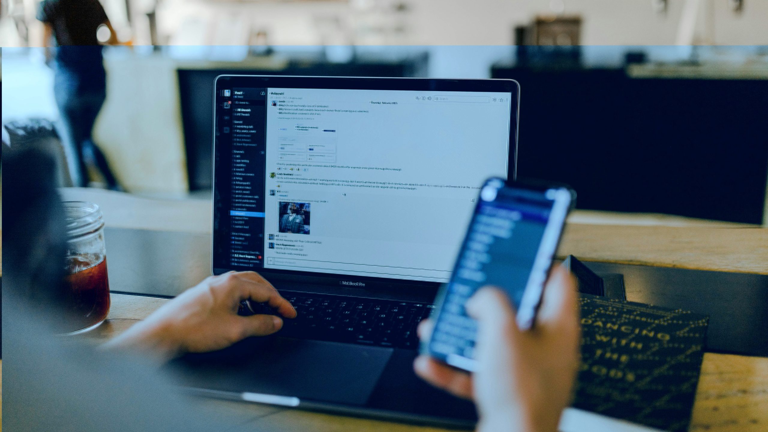 Twee handen met mobiel en op de achtergrond een laptop met wit scherm